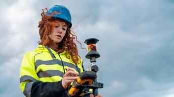 Person mit Helm in Schutzbekleidung steht unter blauem Himmel, Wind weht durch Haar und blickt auf Messgerät, das sie in Händen hält.