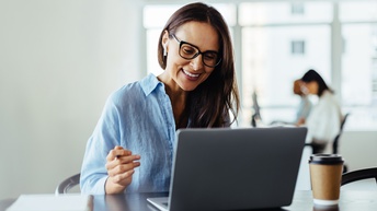 Nahaufnahme einer Person mit Brille, die vor einem aufgeklappten Laptop sitzt. Der Laptop steht auf einem dunkelbraunen Holztisch, daneben steht ein Coffee-to-go-Becher, ebenso liegen ein Notizblock und ein Smartphone auf dem Tisch