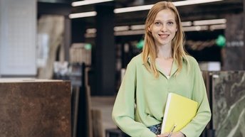 Lächelnde Person im Portrait mit gelber Mappe und Bleistift in Händen, im Hintergrund verschwommen Steinplattenmuster