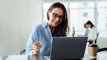 Nahaufnahme einer Person mit Brille, die vor einem aufgeklappten Laptop sitzt. Der Laptop steht auf einem dunkelbraunen Holztisch, daneben steht ein Coffee-to-go-Becher, ebenso liegen ein Notizblock und ein Smartphone auf dem Tisch