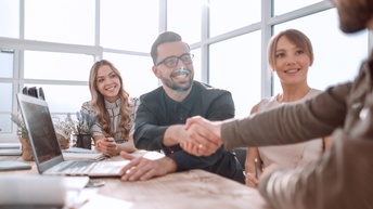 Mehrere lächelnde Personen sitzen an Tisch mit Laptop und Dokumenten, zwei Personen reichen sich die Hände