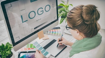 Person sitzt vor einem PC und zeichnet ein Logo während sie eine Farbkarte in der Hand hält