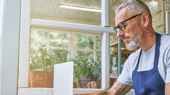 Person mit kurzen grauen Haaren, Bart, Tattoos und Brille trägt eine blaue Schürze und sitzt bei einem Gartentisch mit einem aufgeklappten Laptop, dahinter zeigt sich ein Fenster eines Glashauses mit Pflanzen