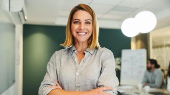 Halbporträt einer Person in einem grauen Hemd und mit verschränkten Armen in einem Raum stehend. Hinter ihr ist ein langer Tisch, an dem zwei Personen sitzen sowie ein Flipchart. Auf dem Tisch liegen Unterlagen.
