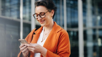 Porträt einer Person mit Brille, orangem Mantel und weißer Bluse steht vor einem Gebäude. Sie blickt in ein Smartphone, das sie mit beiden Händen hält