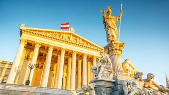 In einem Brunnen ist ein Sockel. Auf diesem sitzen rundherum verschiedene Statuen. In der Mitte ist eine Säule. Auf dieser steht eine weitere große Statue. Hinter dem Brunnen ist ein großes, historisches Gebäude mit einem tempelartigen Teil mit Säulen