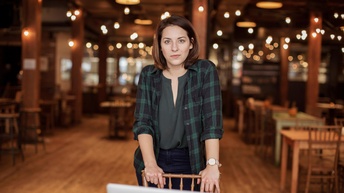 Portrait von einer Person mit schulterlangen braunen Haaren, grüner Bluse und grünem kariertem Hemd vor einem beleuchteten Restaurantambiente.