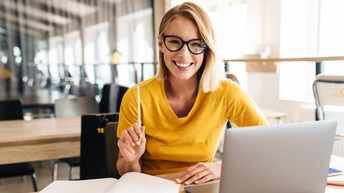 Lächelnde Person mit Brille und erhobenem Stift in der Hand sitzt vor aufgeklapptem Laptop und Büchern 