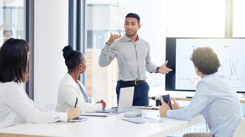 An einem Tisch sitzen drei Personen in Bürokleidung. Neben dem Tisch steht eine weitere Person, die zu den anderen spricht. Neben ihr ist ein großer Bildschirm, auf dem Statistikkurven sind