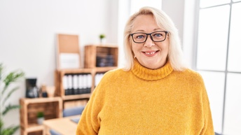 Porträt einer lächelnden Person mit Brillen und gelbem Pullover, im Hintergrund verschwommen Besprechungszimmer mit Regalen