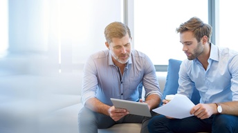 Zwei Personen in hellblauen Hemden sitzen auf Polstermöbel und blicken auf Tablet, eine Person hält das Tablet, die andere hat ein Blatt Papier und einen Kaffeebecher in Händen