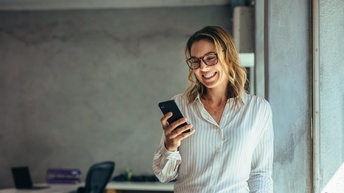 Person mit Brillen blickt freudestrahlend auf Smartphone, im Hintergrund verschwommen Schreibtisch mit Sessel