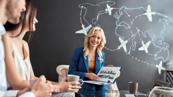 In der linken Bildhälfte sitzen zwei Personen an einem Tisch. Neben ihnen sitzt eine weitere Person, die einen aufgeklappten Ordner hält, auf dem Best Places steht. Hinter dieser Person ist eine dunkle Wand, auf der die Kontinente der Welt sind