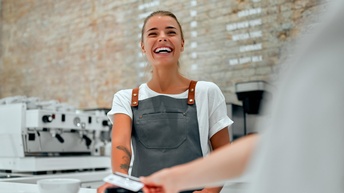 Frontale Aufnahme einer lächelnden Person in Schürze, die hinter einem Tresen steht und einer anderen Person ein Bankomatlesegerät hinhält. Die andere Person hält ihre Hand auf das Gerät. Im Hintergrund ist eine Espressomaschine
