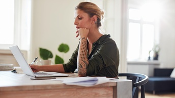 Person mit zusammengebundenen blond-braunen Haaren und Tattoos auf den Händen sitzt mit einem grünen Blusenkleid an einem Schreibtisch und notiert sich etwas während daneben ein Laptop, ein Smartphone sowie Unterlagen liegen