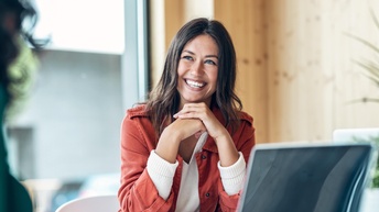 Frontale Aufnahme einer lächelnden Person mit offenen, dunklen Haaren, die an einem Tisch sitzt. Ihr gegenüber sitzt eine weitere Person