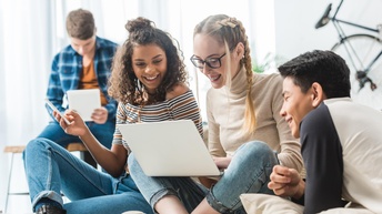 Drei lachende junge Personen auf Couch sitzend und auf Laptop blickend, im Hintergrund weitere Person auf Tisch sitzend