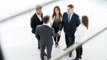 Fünf Personen in Businesskleidung stehen in einem Kreis, zwei der Personen geben sich die Hand