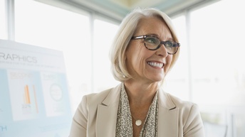 Porträt einer lächelnden Person mit Brillen, im Hintergrund verschwommen Flipchart mit statistischen Grafiken und Fenster