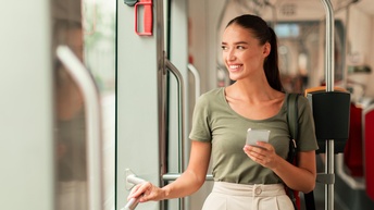 Frontalaufnahme einer Person, die links aus einem Fenster blickt und in der linken Hand ein Smartphone hält. Sie steht in einem Abteil, in dem hinter ihr Sitze sind sowie Metallstangen um sich festzuhalten