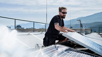 Ein Mann mit Sonnenbrille installiert Photovoltaikanlagen auf einem Flachdach. Im Hintergrund bergige Landschaften.
