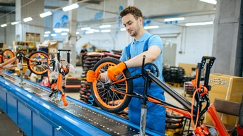 Person in blauer Latzhose mit orangen Handschuhen in Fabrik an Fließband stehend und Räderbestandteile fertigend