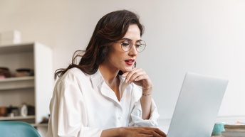 Seitliche Aufnahme einer an einem Tisch sitzenden Person mit Brille, roten Lippen und heller Bluse. Vor ihr steht ein aufgeklappter Laptop, auf dem sie ihre rechte Hand hat. Ihre linke Hand ist unter ihrem Kinn