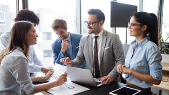 Mehrere Personen stehen an einem Tisch mit Lapop, Tablet und Unterlagen und besprechen Statistiken