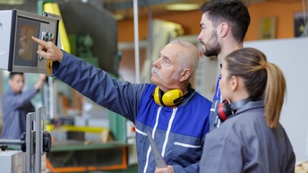 Zwei jüngere Personen in Arbeitskleidung blicken auf Display einer Maschine, auf das ältere Person in Arbeitskleidung mit Hörschutz um den Hals deutet