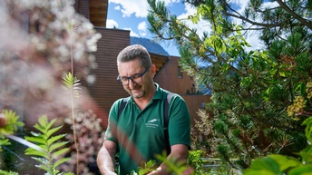 Lächelnde Person mit Brillen steht in Garten und schneidet Pflanzen, im Hintergrund verschwommen Holzfassade, ringsum grüne Pflanzen