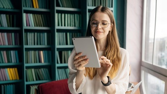 Jugendliche Person mit langen Haaren, Brille und weißer Bluse hält ein Tablet in Händen, im Hintergrund steht eine Couch sowie Regale mit Büchern vor einer offenen hellen Fensterfront