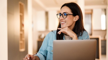 Person mit Brille sitzt vor einem Laptop mit Notizbücher und Stift an einem Tisch und blickt fröhlich und interessiert auf die Seite