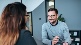 Person mit dunklen kurzen Haaren, Brille und Bart sitzt an einem Tisch und hält ein Klemmbrett sowie ein Stift in Händen während mit einer anderen Person gesprochen wird
