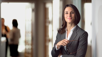 Person mit sanften Lächeln hält Brille in der Hand, im Hintergrund weitere Personen