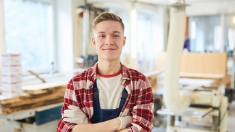 Person mit verschränkten Armen lächelt mild, im Hintergrund Werkstatt mit Maschinen 