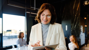 Porträt lächelnden Person mit Brillen, die auf Tablet tippt und blickt, im Hintergrund verschwommen Besprechungsraum mit weiteren Personen