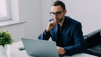 Person mit Brillen blickt auf aufgeklappten Laptop, eine Hand ans Kinn gestützt