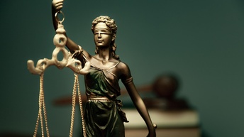 Statue Justitia, im Hintergrund sind Bücher sowie ein Richterhammer in der Unschärfe platziert
