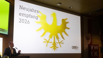 Eine Person steht am Rednerpult und spricht in ein Mikrofon. Hinter der Person ist eine große Leinwand mit der Aufschrift „Neujahrsempfang 2026“ und einem großen goldenen Adler zu sehen. Im Raum sitzen und stehen mehrere Personen an Stehtischen und hören zu. Der Raum ist hell beleuchtet und als Veranstaltungssaal erkennbar.