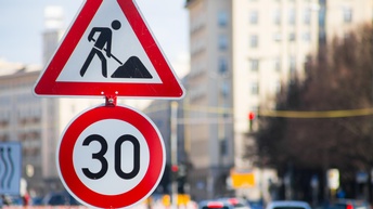 Zwei Straßenschilder im Fokus: Ein Baustellen- und ein 30-er-Zonene-Schild, im Hintergrund verschwommen Autos in Rückansicht.