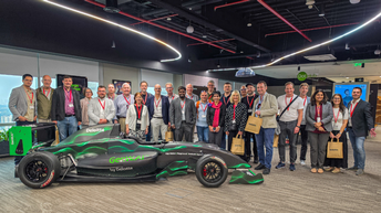 Gruppe von rund 20 Teilnehmer:innen der UBIT-Studienreise steht in einem modernen Büro in Indien neben einem schwarzen Formelrennwagen mit grünen Akzenten („GenW.AI by Deloitte“). 