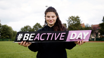 Person hält ein Schild mit der Aufschrift „#BEACTIVE DAY“ auf einer Wiese.