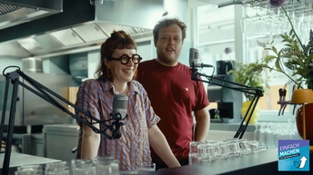 Frontale Aufnahme von zwei Personen, die hinter einem Tresen stehen. Vor den Personen sind zwei Mikrofone angebracht. Hinter ihnen ist eine Küche