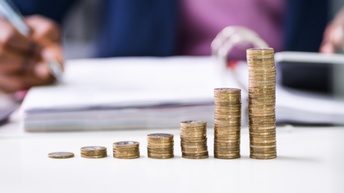  Münzen sind verschieden hoch gestapelt, im Hintergrund sitzt eine Person in der Unschärfe und notiert etwas in einer Mappe mit Unterlagen sowie tippt in einen Taschenrechner, daneben steht ein Laptop