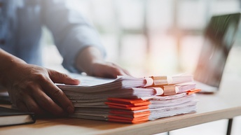 Eine Person greift nach einem Papierstapel, der auf einem Tisch liegt. Daneben steht ein Laptop