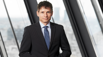 Porträt von Raiffeisen Chefanalyst Gunter Deuber im dunklen Anzug mit dunkelblauer Krawatte, im Hintergrund sieht man verschwommen eine Fensterfront