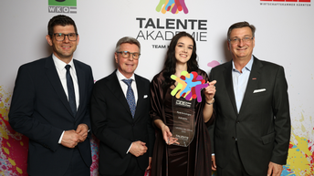 Vier Personen bei der Talenteakademie-Gala der WKO Kärnten; eine junge Frau hält einen Glaspokal, flankiert von drei Männern im Anzug vor einer gebrandeten Eventwand.