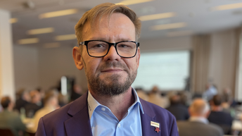 Porträt von einem Mann mit Brille und Businessoutfit - im Hintergrund sieht man verschwommen einen Saal mit Publikum