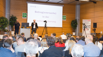 Zwei Männer stehen auf einer Bühne, das Publikum blickt in Richtung Bühne - im Hintergrund auf der Bühne sieht man eine Leinwand mit dem Schriftzug Betrugsversuche in der Sachversicherung
