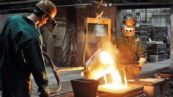 Drei Personen mit Helmen und Schutzkleidung gießen glühendes, flüssiges Metall aus einem Kessel in eine Form